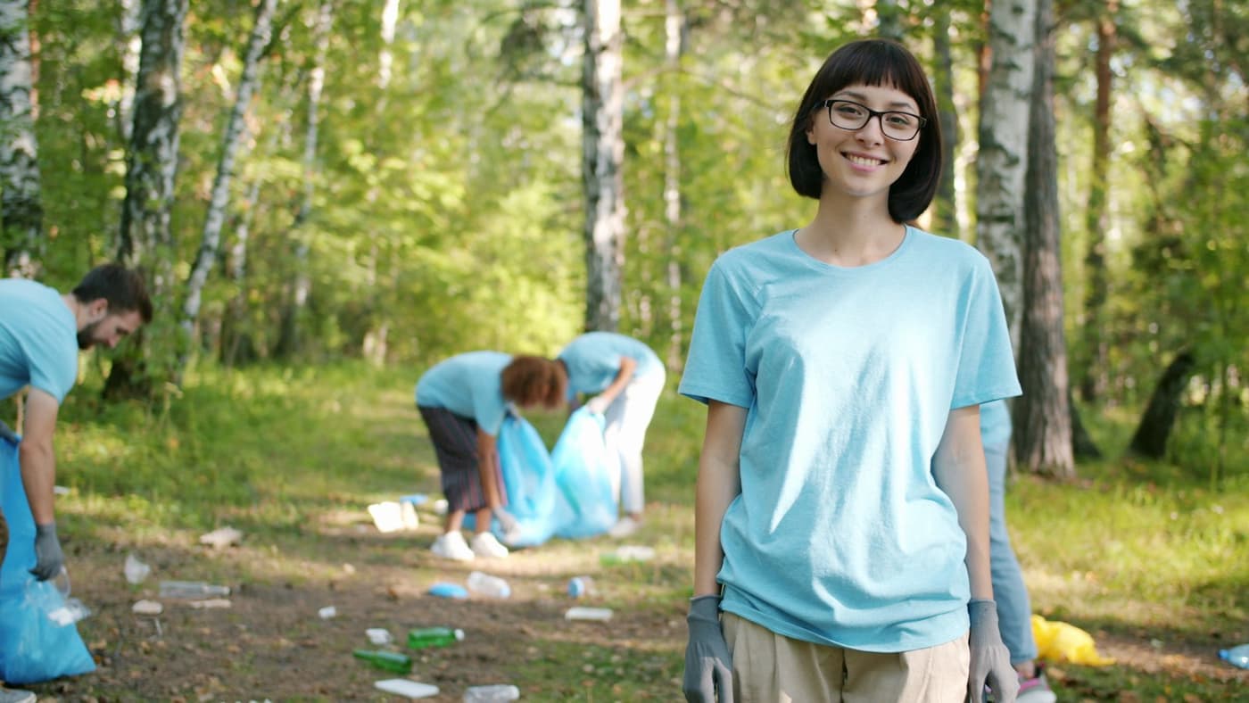A volunteer smiling at the camera while others collect litter in a park behind her